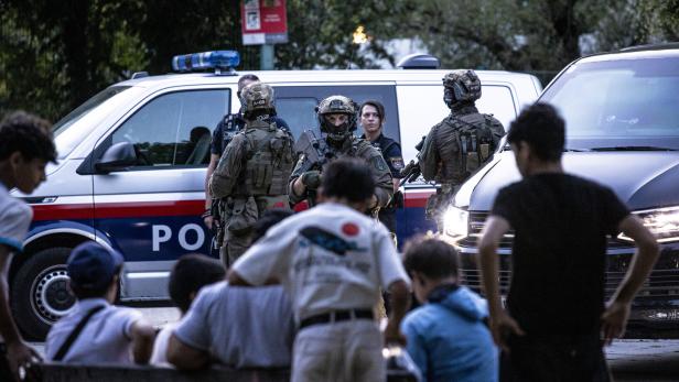 Polizisten in Kampfmontur stehen vor einem Polizeiwagen, während Jugendliche zusehen.