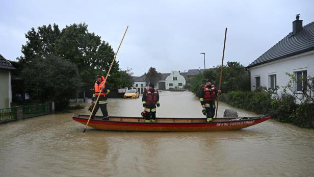Drei Feuerwehrleute in Schwimmwesten navigieren ein Boot durch eine überschwemmte Straße in Gänserndorf.