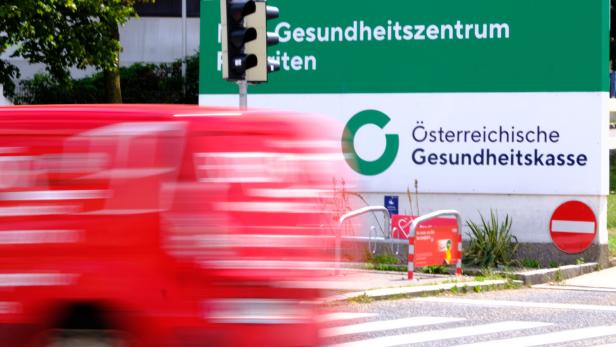 Ein Schild der Österreichischen Gesundheitskasse (ÖGK) mit einem vorbeifahrenden roten Lieferwagen.