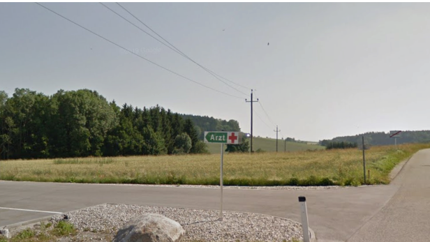 Ein Wegweiser mit der Aufschrift „Arzt“ steht an einer Landstraße vor Feldern und Wiesen am Rand von St. Marienkirchen am Hausruck.