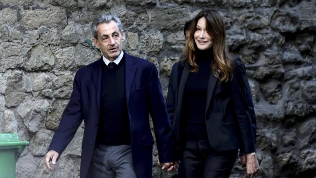 Nicolas Sarkozy und Carla Bruni gehen händchenhaltend vor einer Steinmauer entlang.