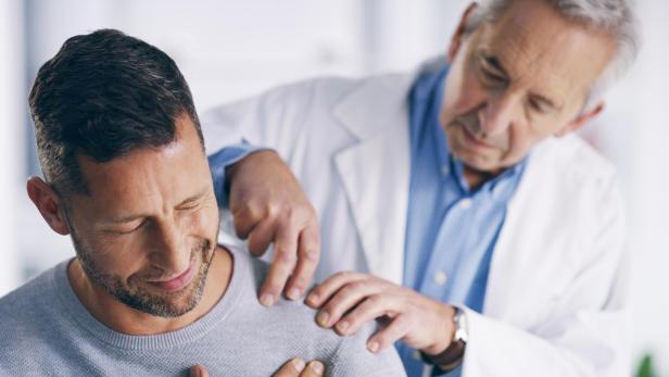 Das Bild zeigt eine Behandlungsszene, bei der ein Arzt einen Patienten untersucht, dessen Schulter schmerzt.
