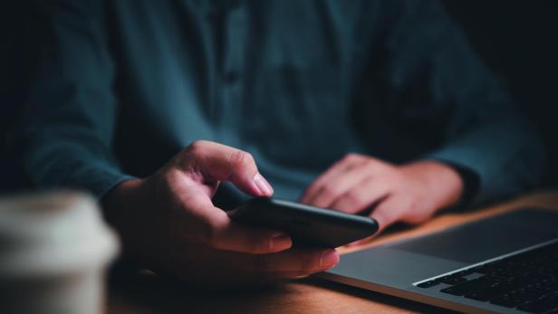 Hand hält Smartphone über einem Holztisch, im Hintergrund ein Laptop und eine Kaffeetasse unscharf sichtbar.