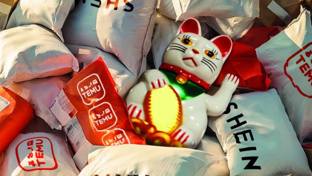 Ein Haufen Plastikbeutel von Shein und Temu, dazwischen liegt eine chinesische Winkekatze.
