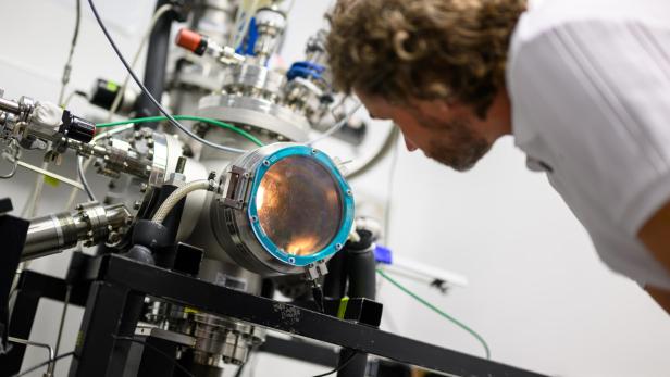 Person mit lockigem Haar beugt sich über ein technisches Gerät mit rundem Sichtfenster und blauen Schrauben in einem Labor.