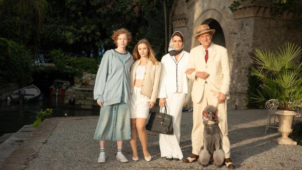 Vier Personen in eleganter Kleidung stehen nebeneinander auf einem Kiesweg vor einem historischen Gebäude, eine Person hält die Leine eines grauen Pudels.