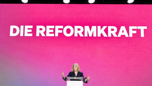 Eine Frau, es ist Neos-Chefin Meinl-Reisinger, spricht am Rednerpult von NEOS vor einem pinkfarbenen Hintergrund mit der Aufschrift "DIE REFORMKRAFT".
