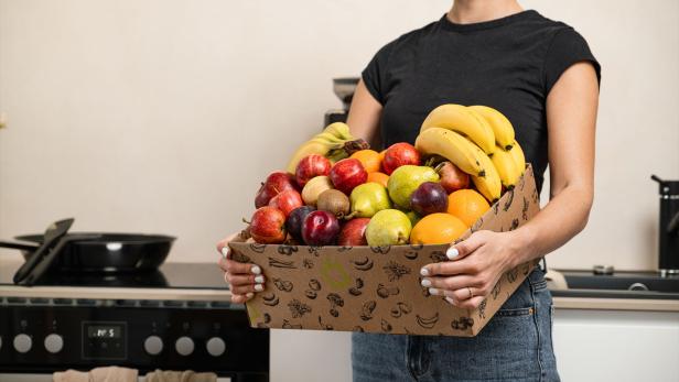 Eine frau in einem schwaren T-Shirt hält einen Obstkorb in den Händen und steht in einer Küche. 