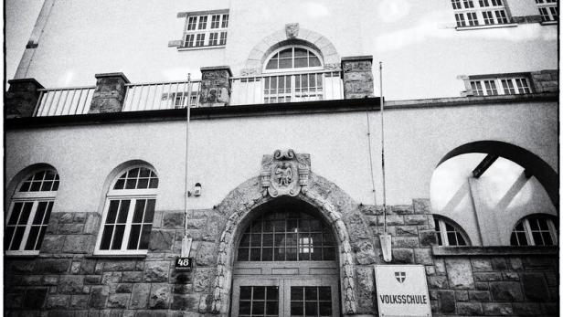 Historisches Schulgebäude mit steinernem Eingangsportal, großen Fenstern und einem Schild mit der Aufschrift „Volksschule“.