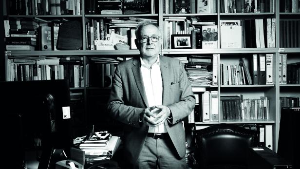 Ein Mann mit Brille und Anzug, es ist der Historiker Oliver Rathkolb, steht vor einem Bücherregal in Schwarzweiß.