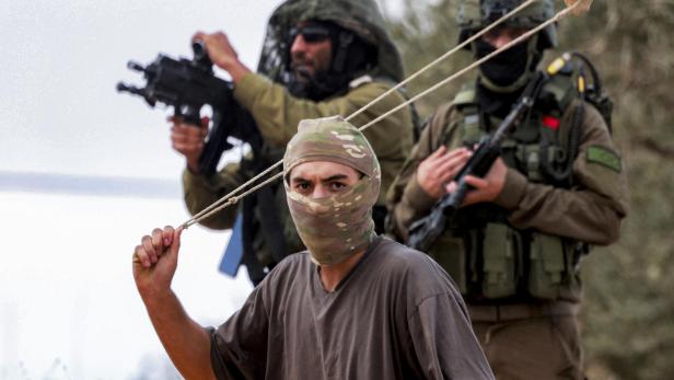 Ein israelischer Siedler steht vor zwei israelischen Soldaten, er lässt eine Steinschleuder über seinen Kopf kreisen.