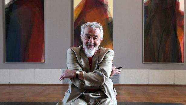 Der Künstler Arnulf Rainer, lachend, in hellem Anzug, hinter ihm drei seiner Übermalungs-Werke