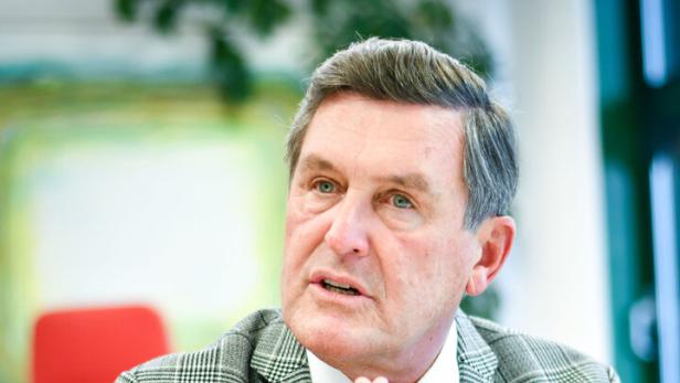 Ein Mann mit kariertem Sakko sitzt mit gefalteten Händen an einem Tisch.