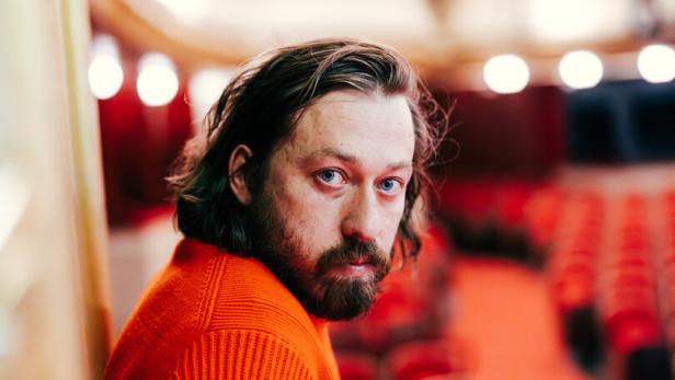 regisseur simon stone im parkett des burtheaters, mit rotem pullover und langen haaren