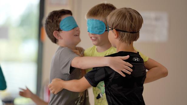 Drei Kinder mit Augenbinden umarmen sich und lächeln dabei.