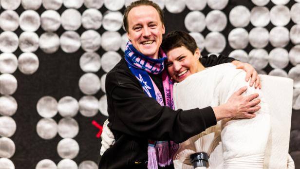 Regisseur Jan-Christoph Gockel und Schauspielerin Alicia Aumüller umarmen einander lachend vor einem weißen Paillettenvorhang.