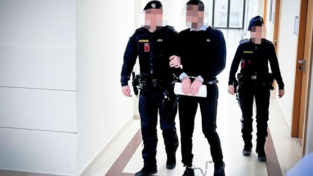 Ein Mann in Handschellen und Fußfesseln wird von zwei Polizisten durch einen Flur geführt.
