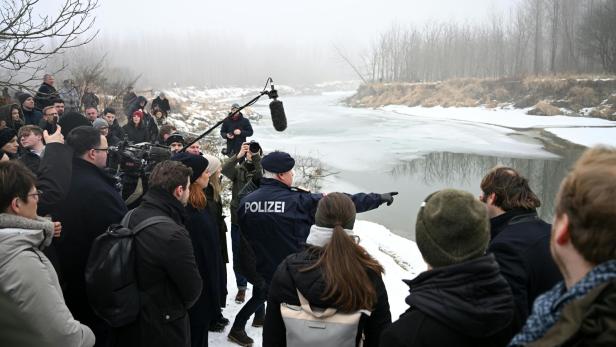 Eine Gruppe von Menschen steht am verschneiten Flussufer, ein Polizist zeigt auf das teilweise zugefrorene Wasser.