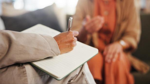 Eine weibliche Hand schreibt in ein leeres Notizbuch, gegenüber sitzt eine andere Frau, die erzählt. Eine Szene aus der Psychothterapie