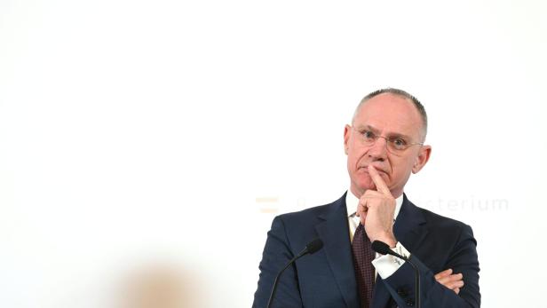 Ein Mann im Anzug (Innenminister Karner) steht nachdenklich mit verschränkten Armen und Finger am Mund vor zwei Mikrofonen.