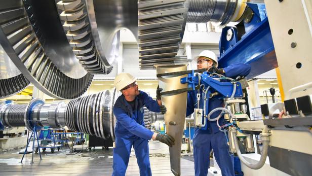 Zwei Arbeiter in blauer Arbeitskleidung und Schutzhelmen montieren ein großes Metallteil in einer Industriehalle mit großen Zahnrädern im Hintergrund
