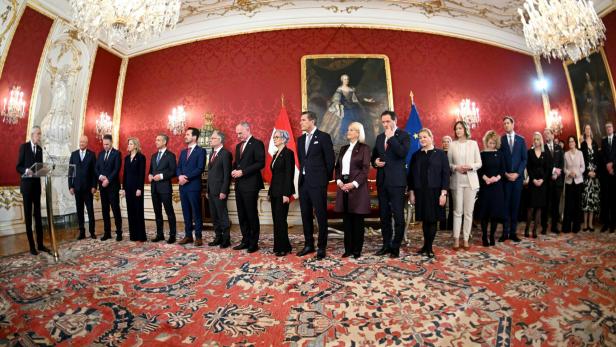 Mitglieder der österreichischen Bundesregierung stehen in einem prunkvollen Saal während einer offiziellen Zeremonie nebeneinander.