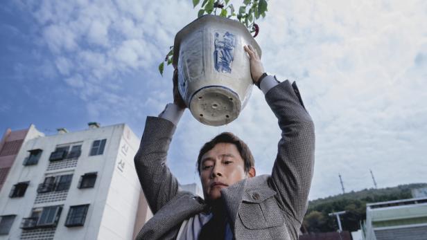 Der Schauspieler Lee Byung-hun, von unten gesehen, mit einem großen Blumentopf, den er wie wurfbereit über seinen Kopf hält 