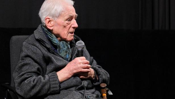 Alexander Kluge spricht in einem schwarz vertäfelten Raum in ein Mikrofon.