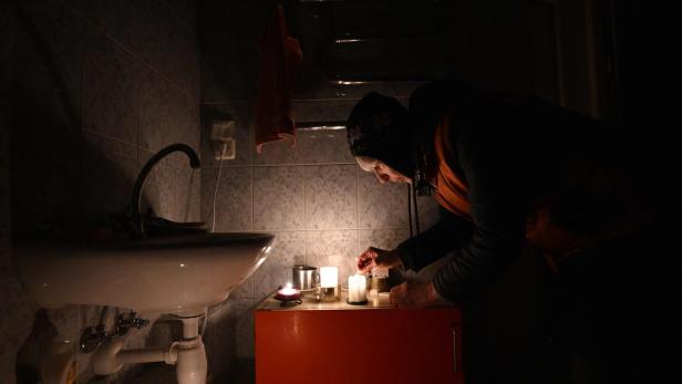 Eine alte Frau, in warme Kleidung und Mütze gehüllt, beugt sich über Kerzen, die sie in ihrem Badezimmer auf einen roten Kasten gestellt hat. Der beflieste Raum ist dunkel. Im Vordergrund ist ein Waschbecken zu sehen.
