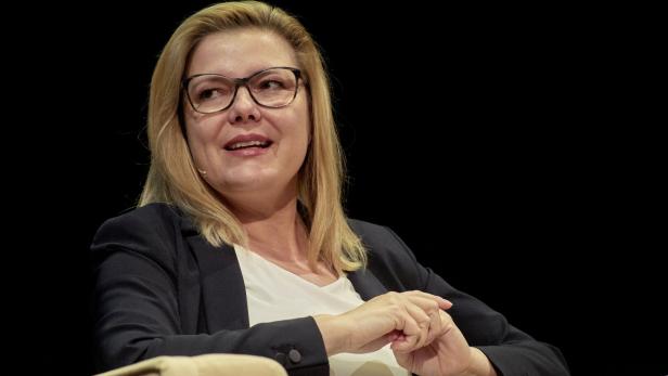 Eine blonde Frau mit Brille und dunklem Blazer sitzt vor schwarzem Hintergrund.