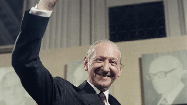 Ein lächelnder Mann im Anzug, Kurt Waldheim im Jahr 1986, hebt den Arm vor einem hellen Hintergrund.