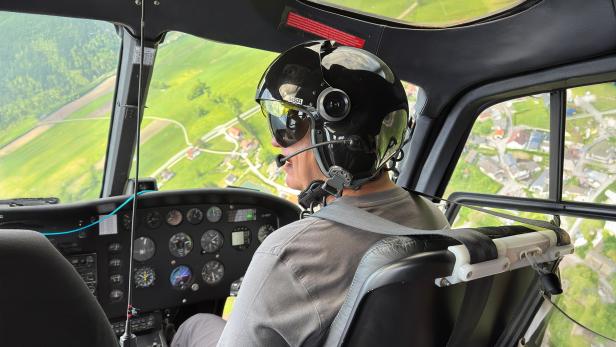 Pilot mit Helm steuert einen Helikopter über eine grüne Landschaft in Österreich.