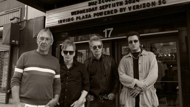 Die vierköpfige Band Buzzcocks in einem schwarzweißen Foto, hinten ein Lokal, das ihr Auftreten annonciert