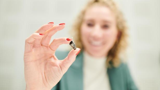 Hand mit roten Fingernägeln hält ein kleines graues Hörgerät zwischen Daumen und Zeigefinger