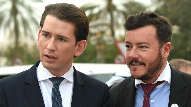 Zwei Männer im Anzug unterhalten sich im Freien, im Hintergrund sind Palmen zu sehen.