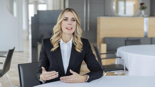 Frau mit langen blonden Haaren in schwarzem Blazer und weißem Hemd sitzt an einem runden Tisch in einem modernen Raum mit Spiegel und Stuhl im Hintergrund