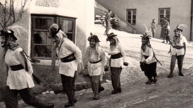 Mehrere Menschen in traditionellen Kostümen mit Masken und Hüten gehen in einer winterlichen Dorfszene hintereinander her.