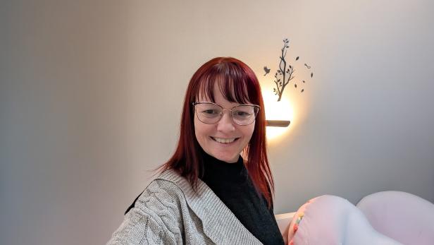 Frau mit Brille und roten Haaren lächelt vor heller Wand mit Wanddeko und Lampe.