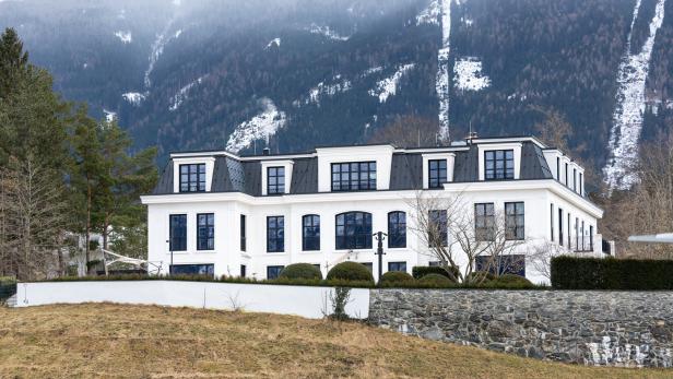 Ein großes, weißes Gebäude mit dunklem Dach steht vor einem bewaldeten Berghang.