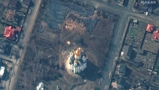Eine Satellitenaufnahme der Stadt Butscha in der Ukraine. Zu sehen ist eine Kirche und auf den Wiesen daneben viele kleine dunkle Objekte - mutmaßlich die Leichen von Zivilist:innen.