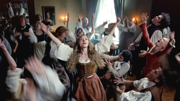 Die Schauspielerin Amanda Seyfried mit ausgebreiteten Armen, umringt von ihrem Ensemble in einem großen Innenraum 