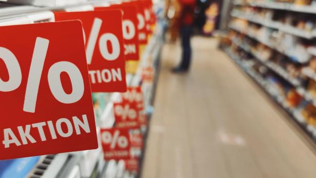 Rote Schilder mit Prozentzeichen und „Aktion“ hängen an einem Supermarktregal, im Hintergrund Regale und eine Person.