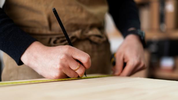 Hände messen und markieren ein Holzbrett mit Bleistift und Maßband in einer Werkstatt.