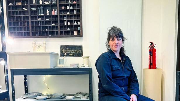 Eine Frau sitzt in einem Atelier auf einem Hocker, umgeben von Regalen mit Keramik und kleinen Figuren.