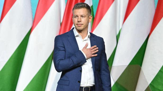 Peter Magyar legt seine Hand auf sein Herz, er steht vor Ungarn-Flaggen.