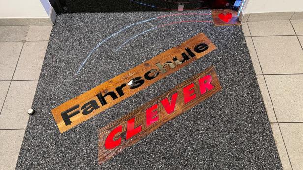 Eine Fußmatte liegt auf einem Fließenboden vor einer Eingangstüre mit der Aufschrift "Fahrschule Clever".