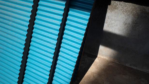 Drei blaue Wellblechplatten lehnen an einer Wand, daneben fällt ein rechteckiger Schatten auf den Boden.