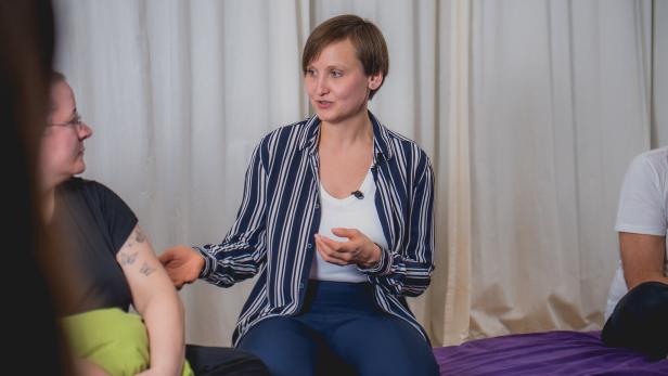 Eine Frau im gestreiften Hemd spricht mit einer anderen Person, beide sitzen entspannt auf lila Decken.