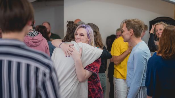 Mehrere Menschen stehen zusammen, zwei Personen umarmen sich herzlich in der Mitte, während andere sich unterhalten.