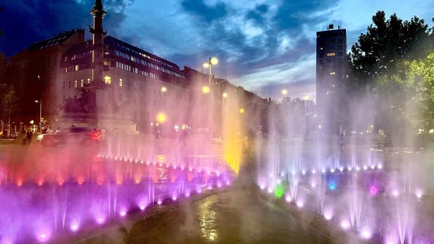 Ein Stadtweg in der Abenddämmerung. Rechts und links vom Weg sprühen bunt beleuchtete Fontänen Wassertröpfchen in die Luft.